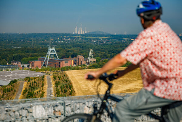 Ein Radfahrer blickt von der Halde Hohewart aus über die Landschaft. Zu sehen sind ein Förderturm sowie eine weitere Halde und industrielle Anlagen am Horizont.
